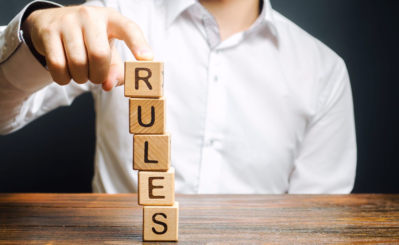 A man holds a tower of blocks with the word Rules from falling. Setting clear rule and restrictions. Leadership and discipline. Authoritarianism, tight control framework. Norms and laws in society